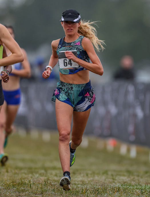Katie Izzo looks determined as she runs in a cross country race, wearing an Adidas uniform and a black hat with a light stripe on either side.