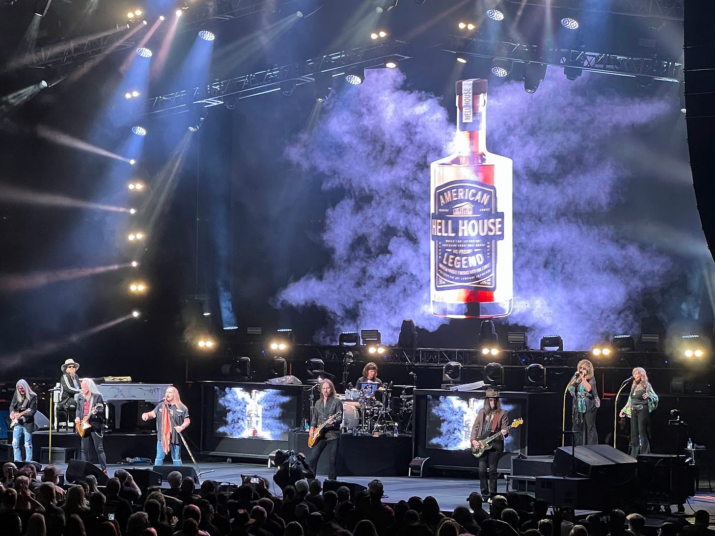 A wide shot cell phone photo of the band Lynyrd Skynyrd playing on stage. Behind the band is a giant video screen displaying a bottle of "Hell House" brand whiskey.