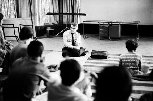 A black-and-white photo of a man in a shirt and tie sitting on the floor in front of a group facing him.