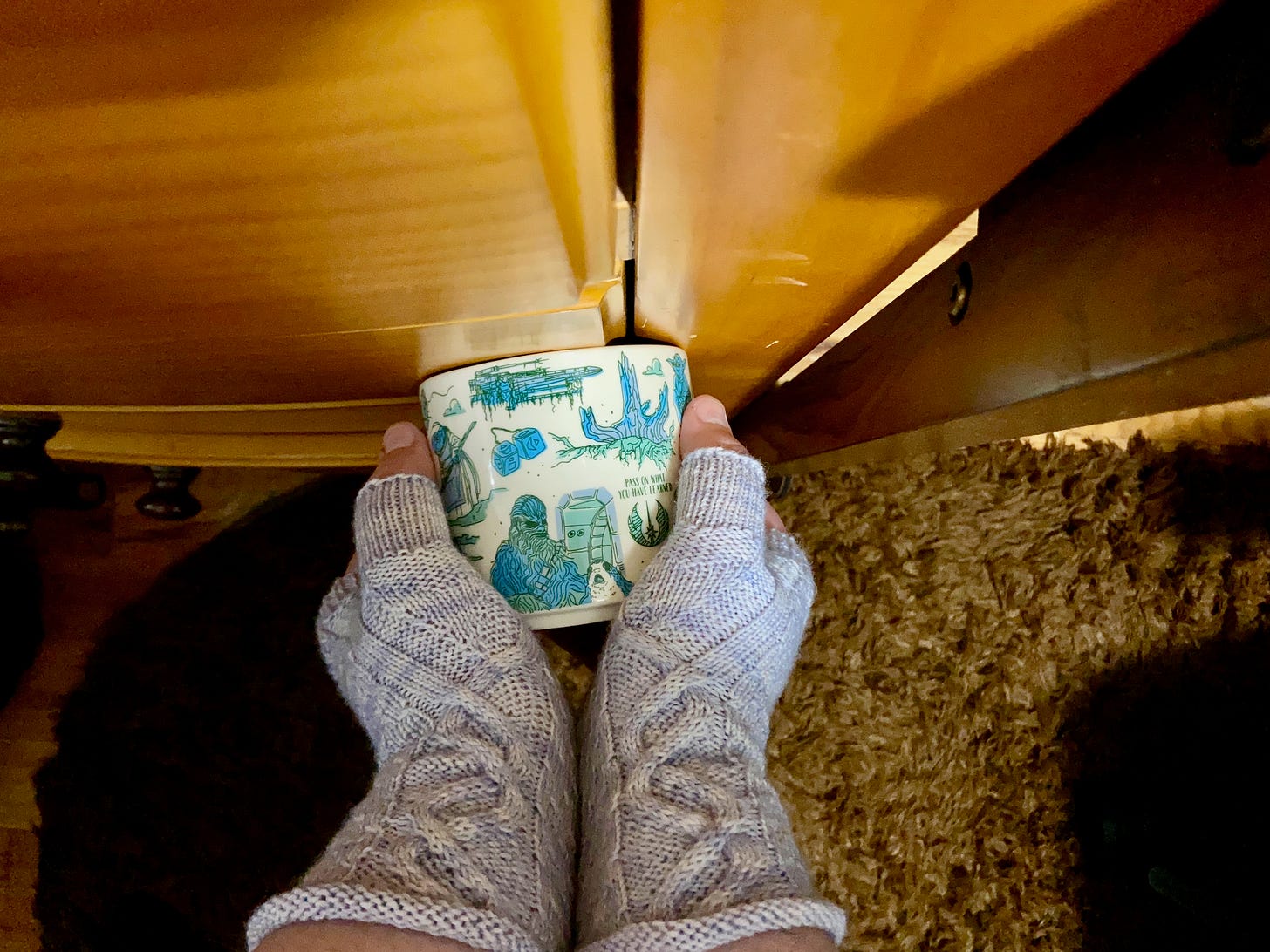 I'm wearing lavender-colored fingerless gloves that I knitted by hand while holding a Star Wars-themed coffee cup sideways. It's positioned against a dresser, and you can see a brown rug below.