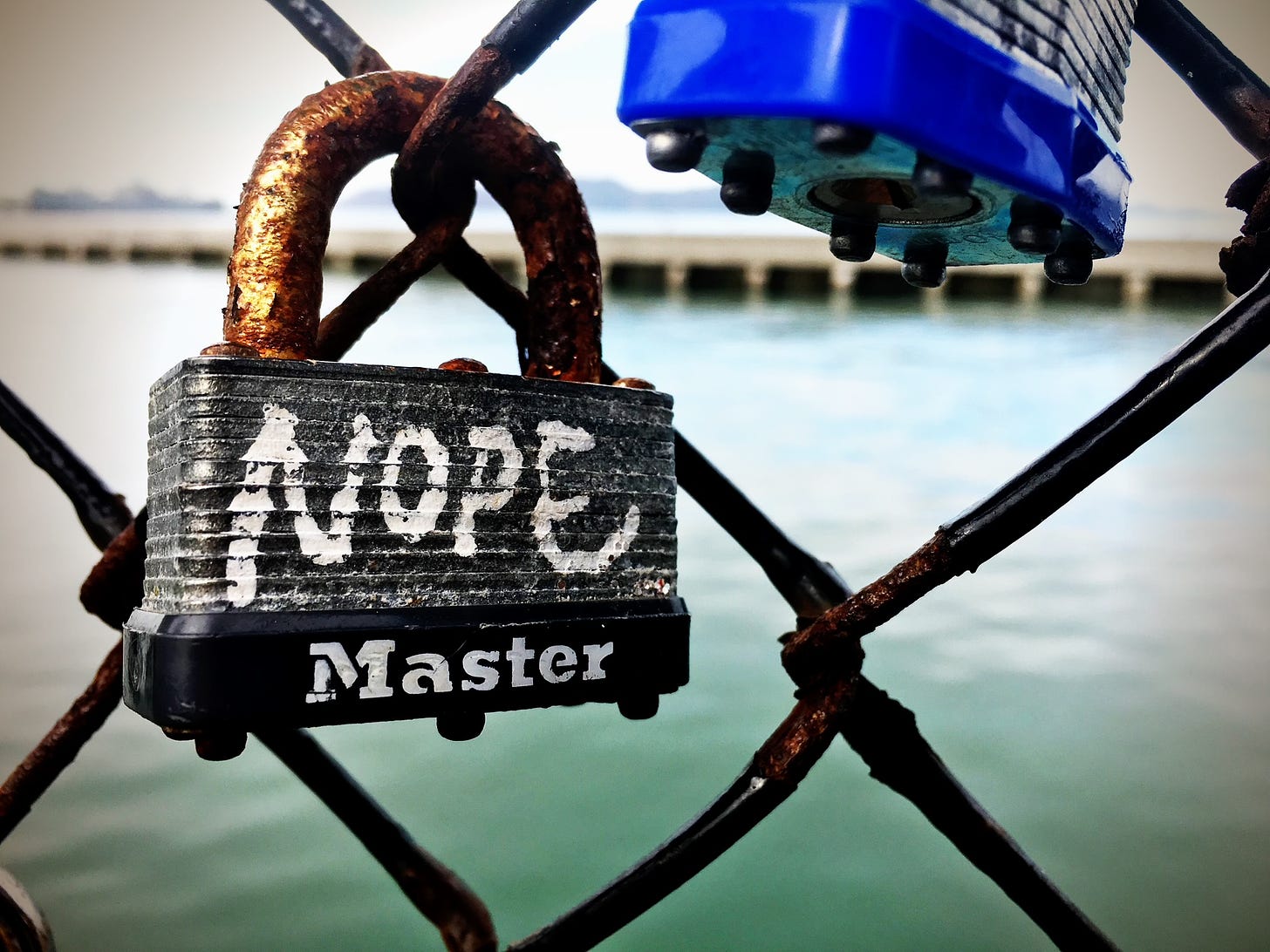 2 padlocks are on a chain-link fence that has as its background the water of a harbor. On one of the locks, the word NOPE has been written.