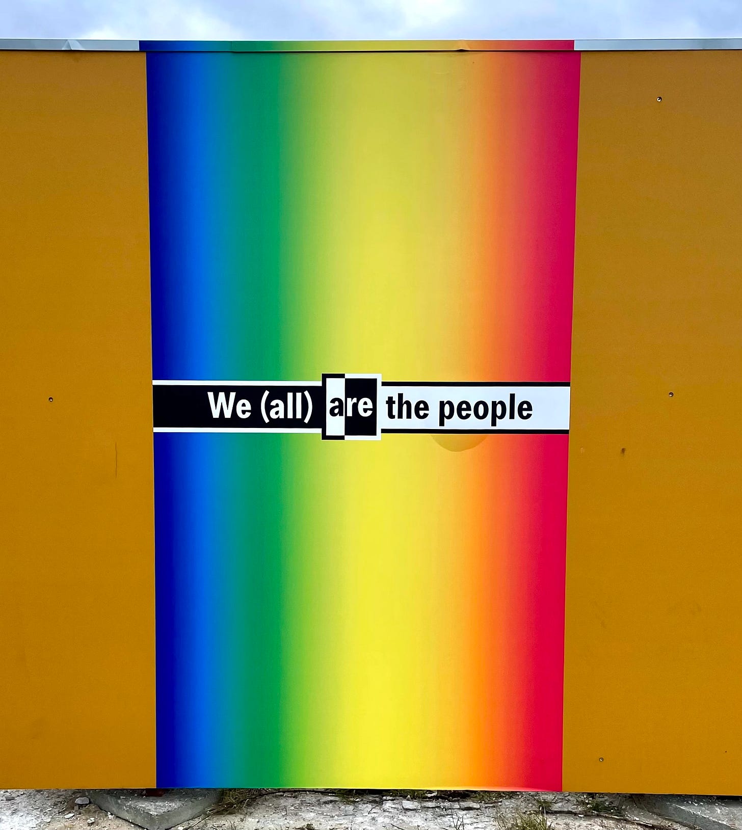 A colorful construction wall in Berlin featuring a gradient rainbow background with the words "We (all) are the people" prominently displayed in black and white text. Photo by Jay Siegmann. A colorful construction wall in Berlin featuring a gradient rainbow background with the words "We (all) are the people" prominently displayed in black and white text. Photo by Jay Siegmann.