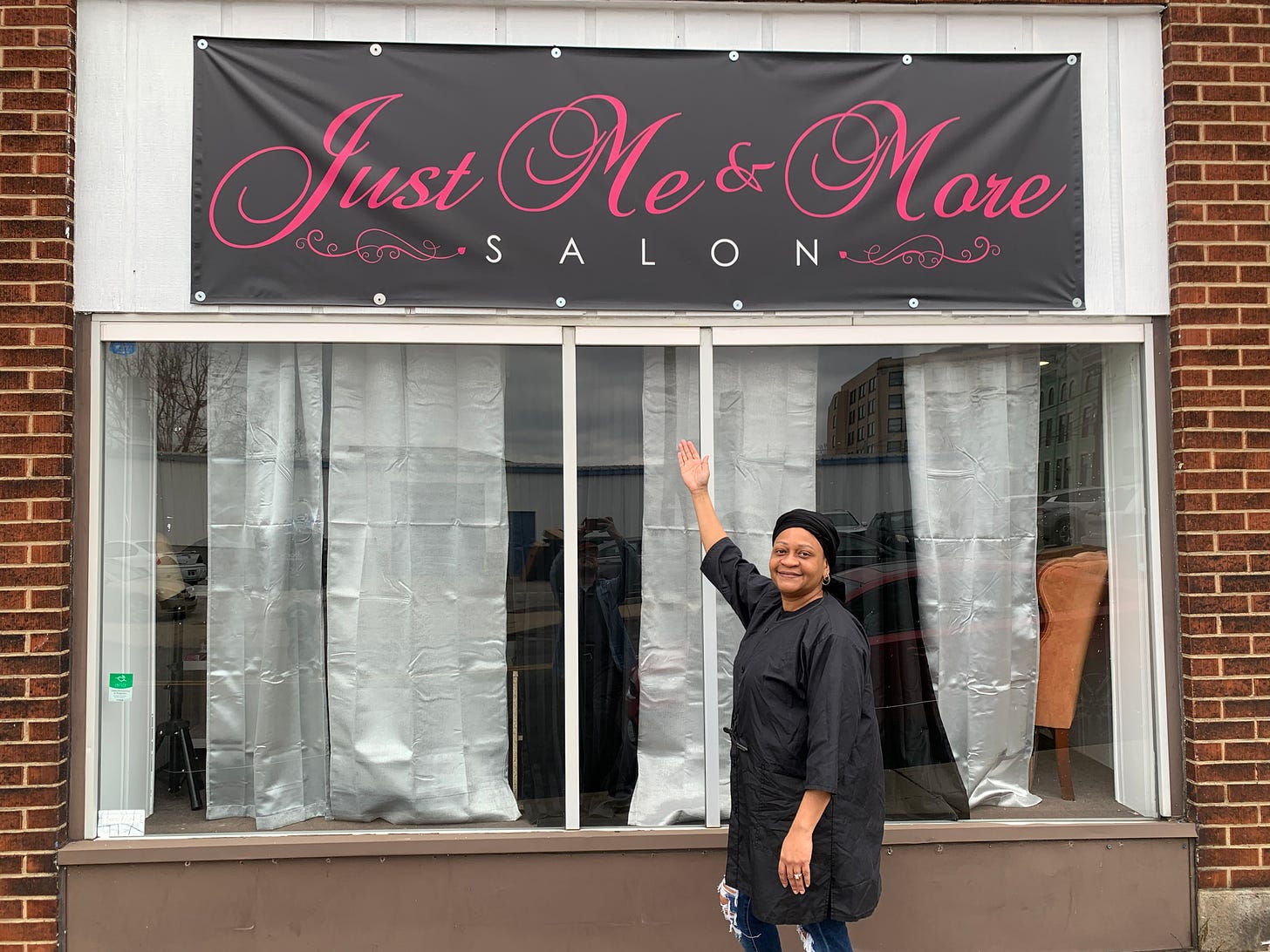 A picture showing Ms Rica Dabney gesturing towards her business' sign that says "Just Me & More" salon