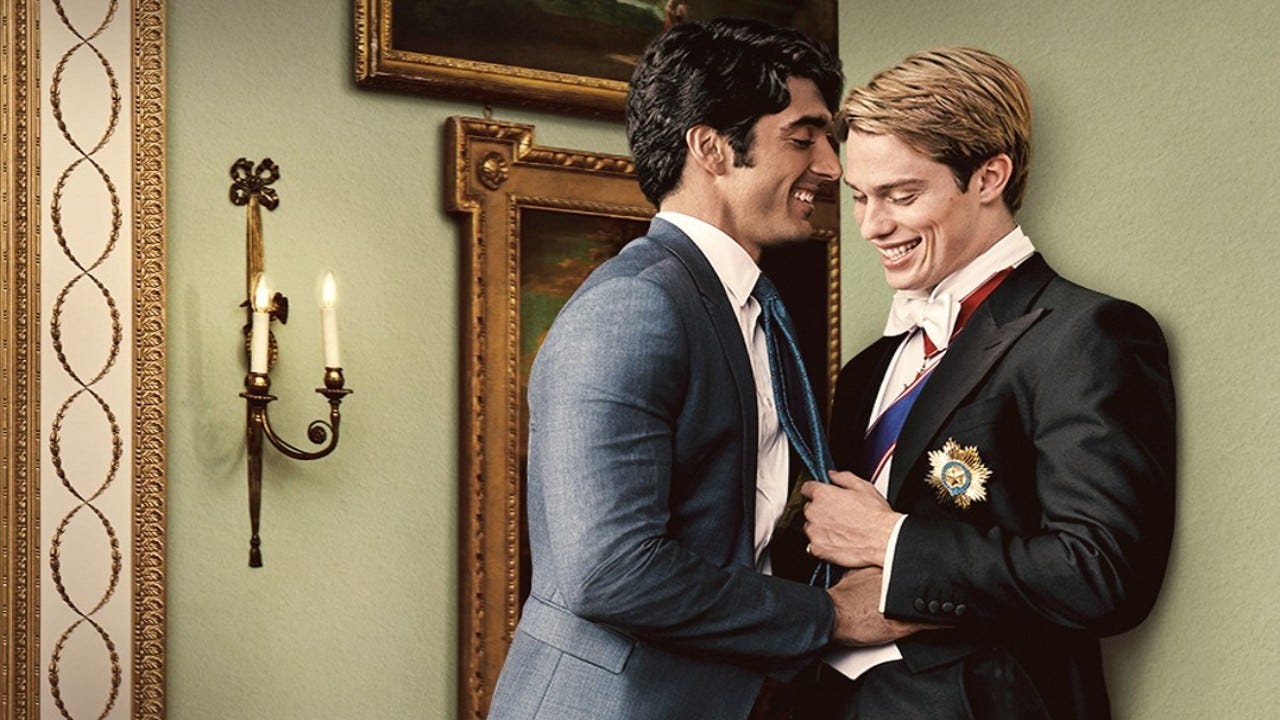 In a formal interior setting with sage green walls and gilded-framed paintings, a Mexican man with short dark hair in a light grey suit presses a blonde white man in a British royal suit against the wall intimately while they both smile big.