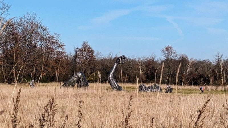 Photo of a large metal sculpture in a grassy field. The sculpture is a person reaching up out of the ground.