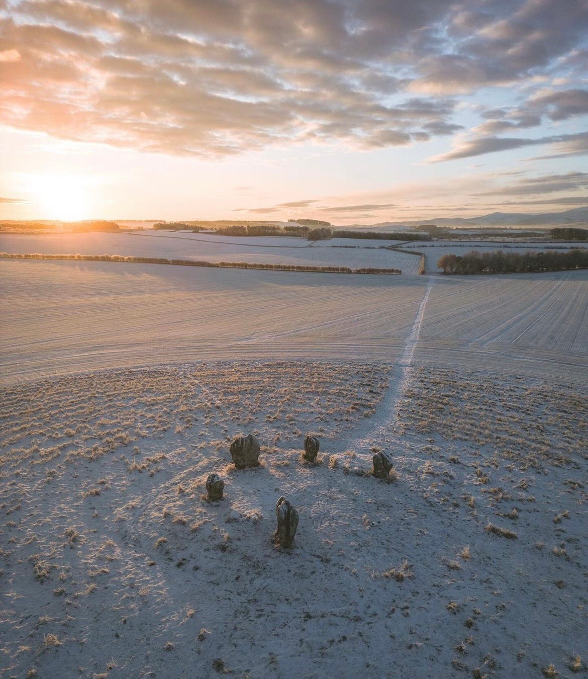 A circle of five stones lies atop a snowy hill in the centre of a field. An open bright but cloudy sky is above them, with the Sun rising to the left of the image.