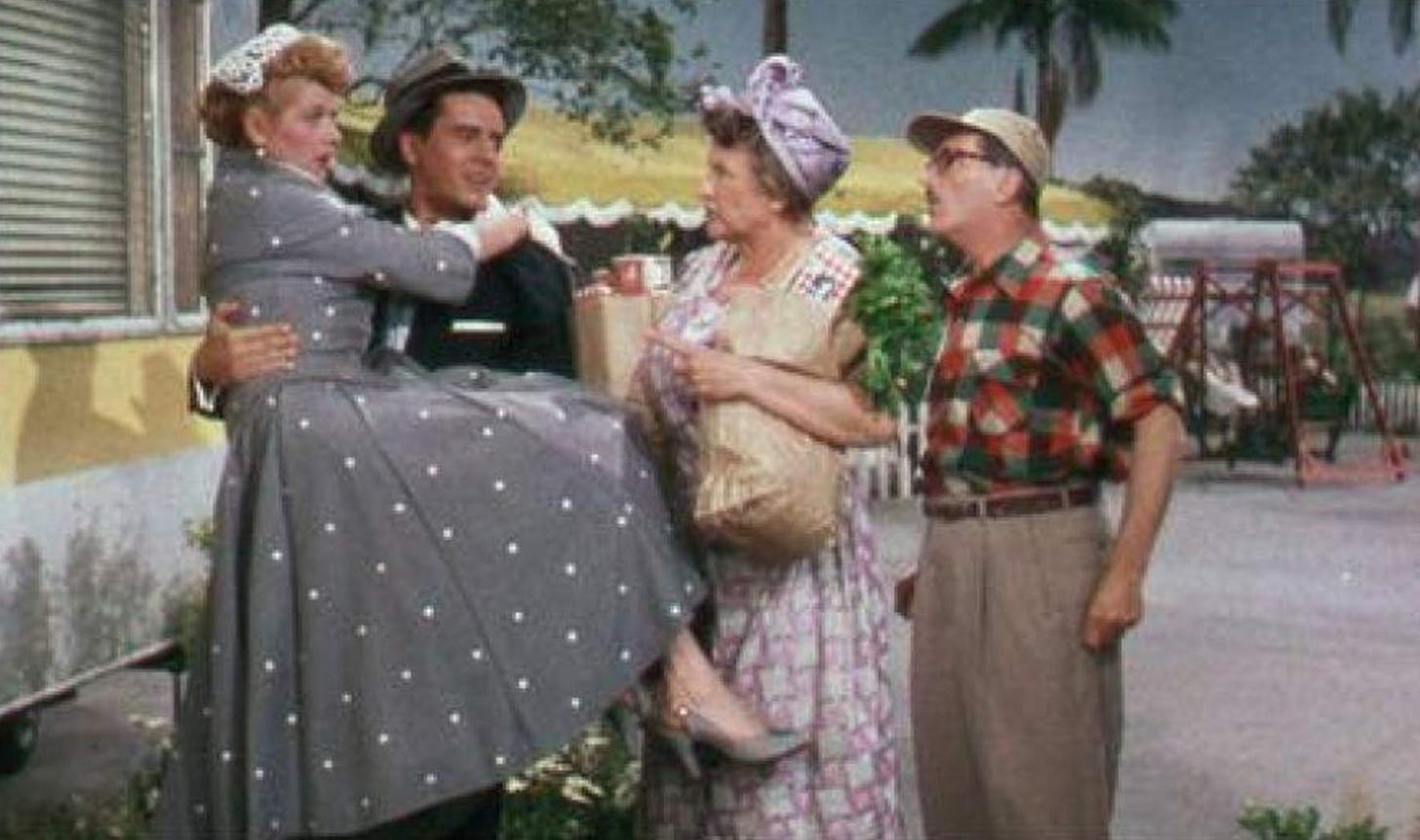 Still from The Long, Long Trailer where Desi Arnaz is carrying Lucille Ball up to the door of their yellow pull-behind trailer, and several neighbors have come up to talk to them.