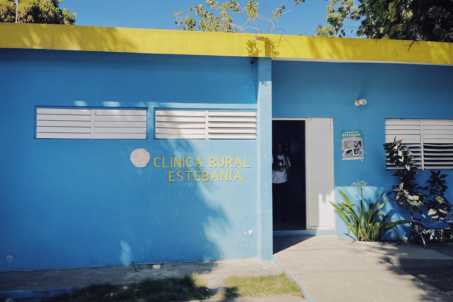 A bright blue building with yellow trip and blue sky visible in the background, with a sign that says CLINICA RURAL ESTEBANIA in yellow letters.
