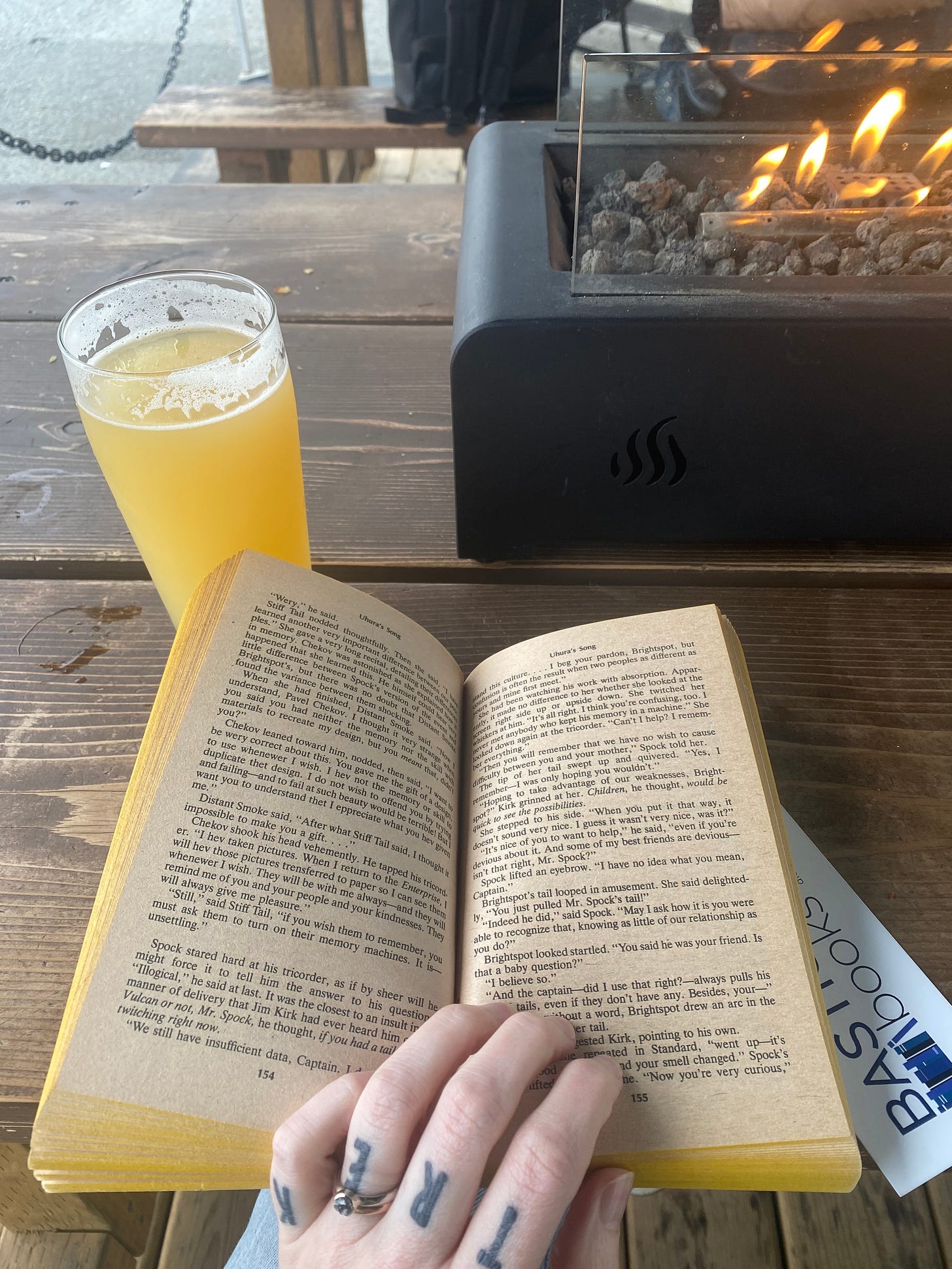 A wooden patio table with a rectangular fire dish with rocks in it. In front of it my hand is holding open a paperback book, "Uhura's Song". Next to the book is a glass of IPA.
