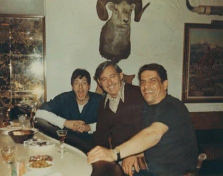 Al Pacino and two other men sitting at a bar with drinks and nibbles in front of them, with paintings and a stag’s head on the wall behind thme