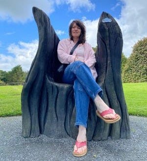 Woman in blue jeans and pink top sitting with legs crossed on a black throne made of bog oak with trees and grass and blue sky in background.