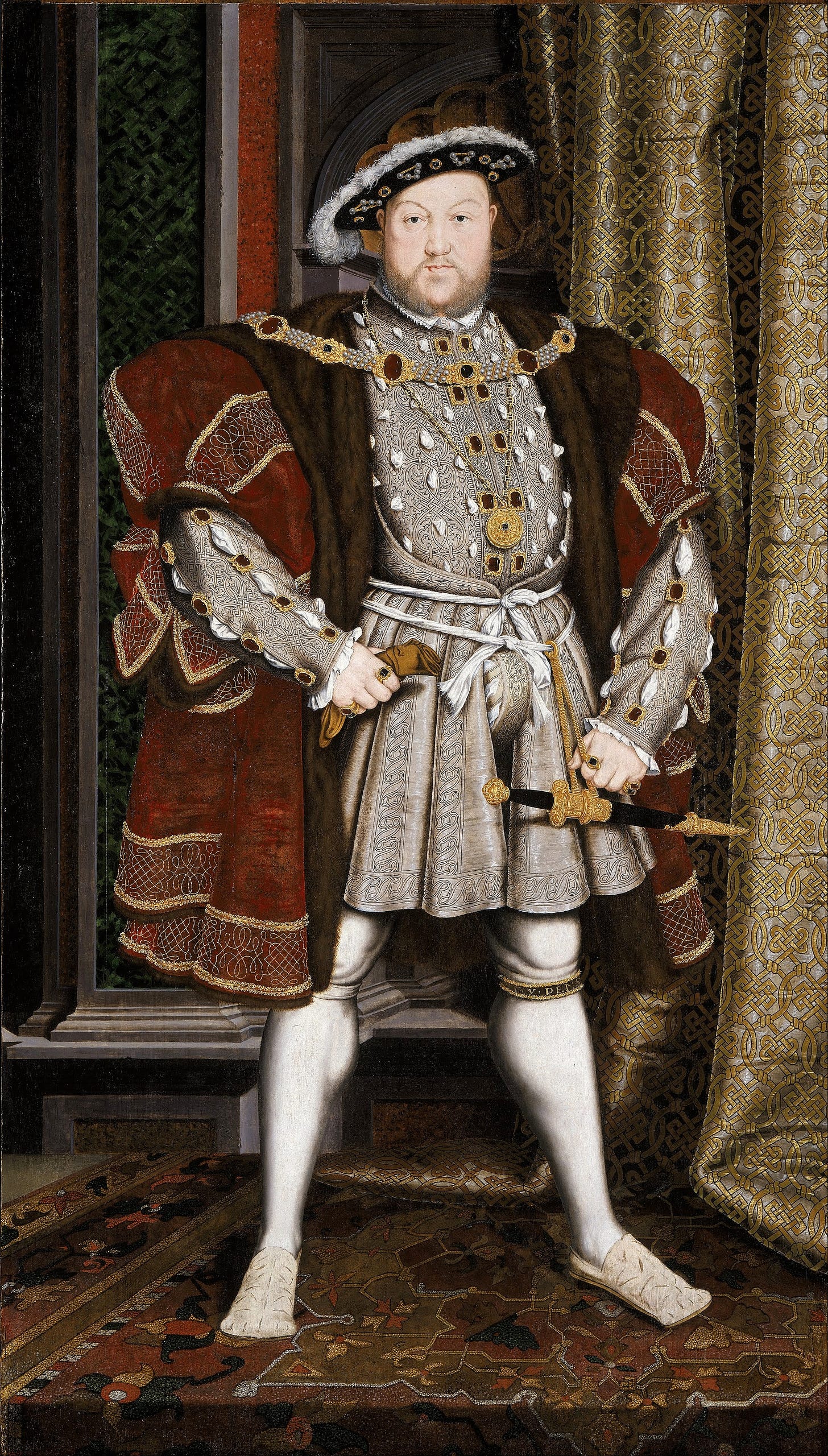 A portrait of King Henry VIII. He stands proudly in front of an ornate background. He wears a silver doublet and a scarlet jacket. He wears other signs of power including a ruby, gold, and pearl chain, a prodding codpiece, and other ornamentation. He holds himself proudly and carries a dagger, clearly a man at the height of his power.