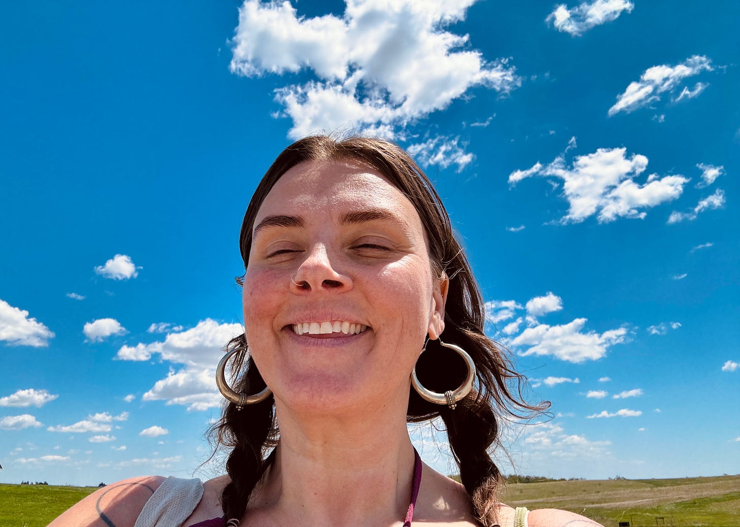 Lisette smiles in the foreground with eyes closed, behind her is a massive blue sky, white clouds and flat green field.