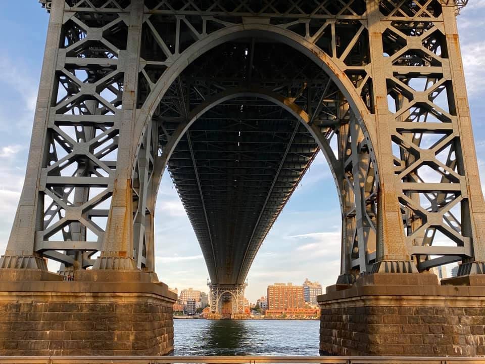 Looking up at the metal underside of the Manhattan Bridge from East River Park. There is blue sky and thin clouds above and the East River below.