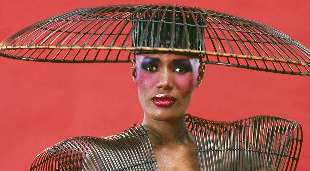 A Black woman, Grace Jones, is wearing jewel-toned makeup and artful cage-structured hat and top, in front of a red background.