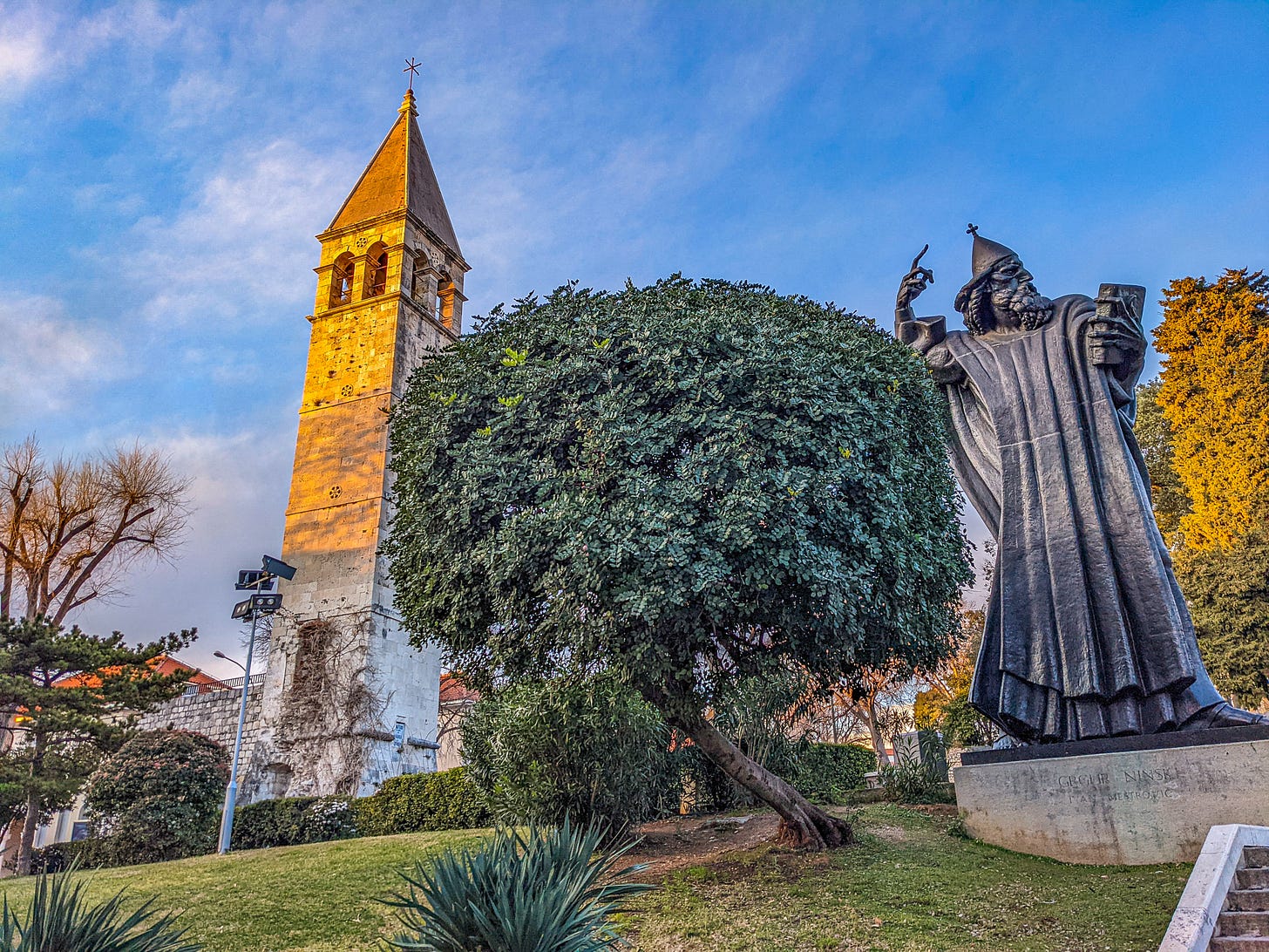 Gregory of Nin standing in a park with trees and a large stone tower behind him. 