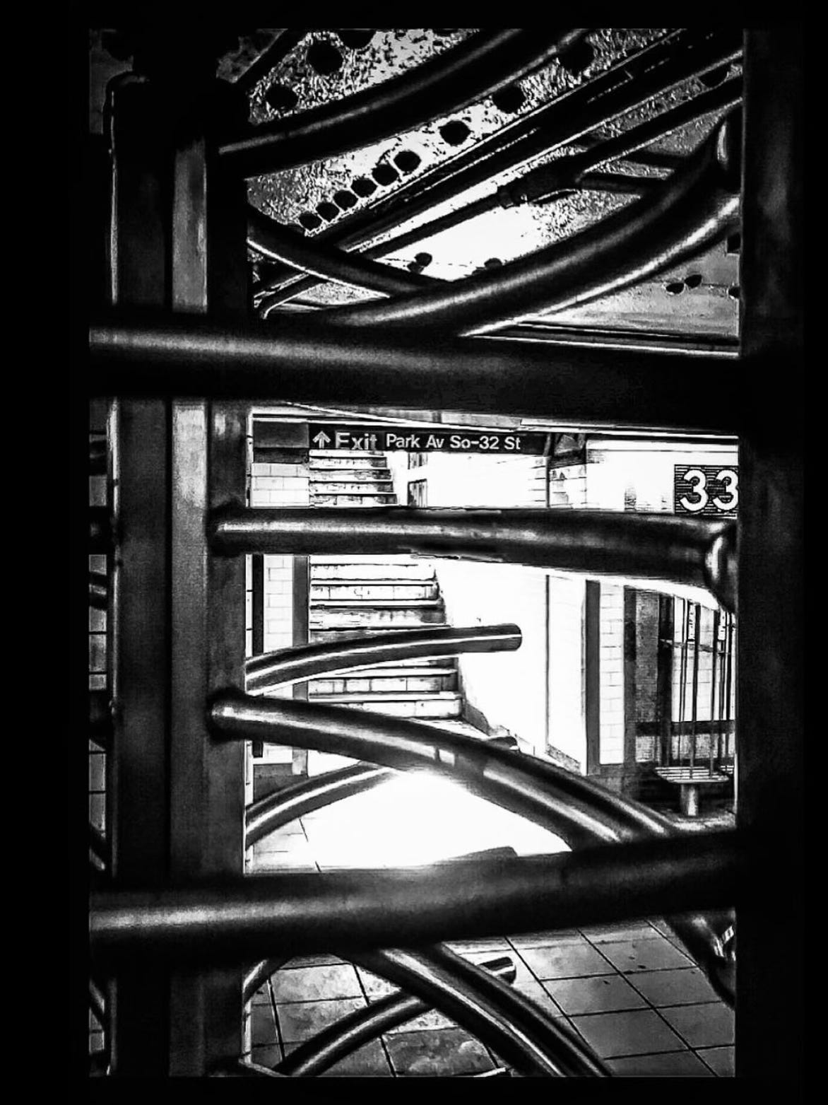 A noir-style black and white photograph of the New York subway, as seen through one of the vertical turnstile entrances. In the foreground, the curving bars of the turnstile make dramatic gestures across the image. Through the bars you can see a stairway with the words “Exit Park Av So-32 St” and on a wall to the right a tile mosaic with the number 33. 