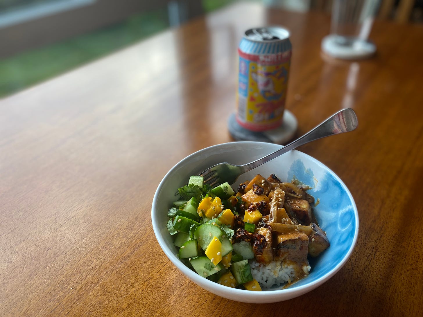 A white and blue bowl of the rice dish described above: red curry tofu and onions on one side, the mango cucumber salad on the other. Chili crisp is drizzled over top, and a can of beer rests on a coaster in the background.