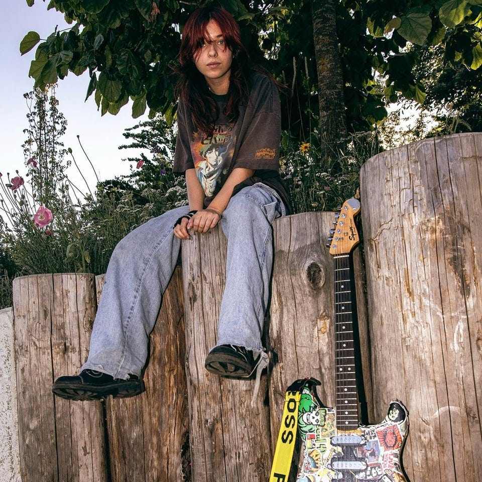 Cherry Pick sitting on a fence near her Squire guitar