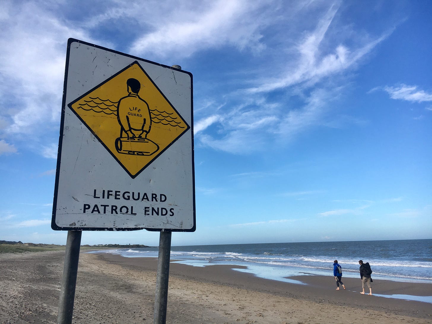 Strand mit blauem Himmel, im Vordergrund ein Schild mit der Aufschrift "Lifeguard Patrol Ends", dahinter laufen zwei Menschen.