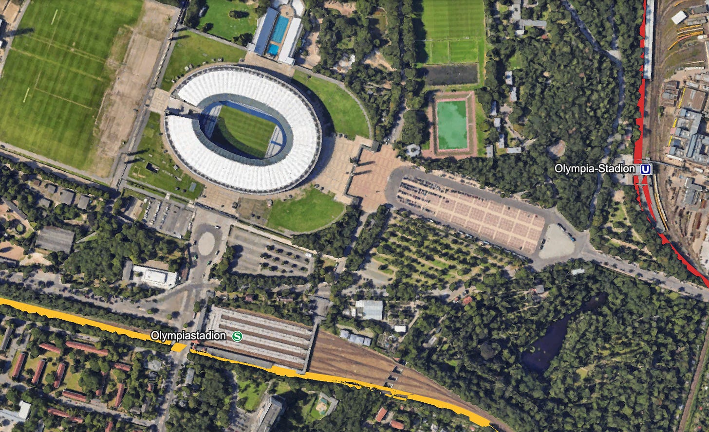 Satellite view of Berlin's Olympiastadion, showing the stadium surrounded by green spaces. The S-Bahn station (Olympiastadion) is marked in yellow and located near the bottom left of the image, while the U-Bahn station (Olympia-Stadion) is marked in red on the right side, illustrating the stadium's accessibility via both transit systems.