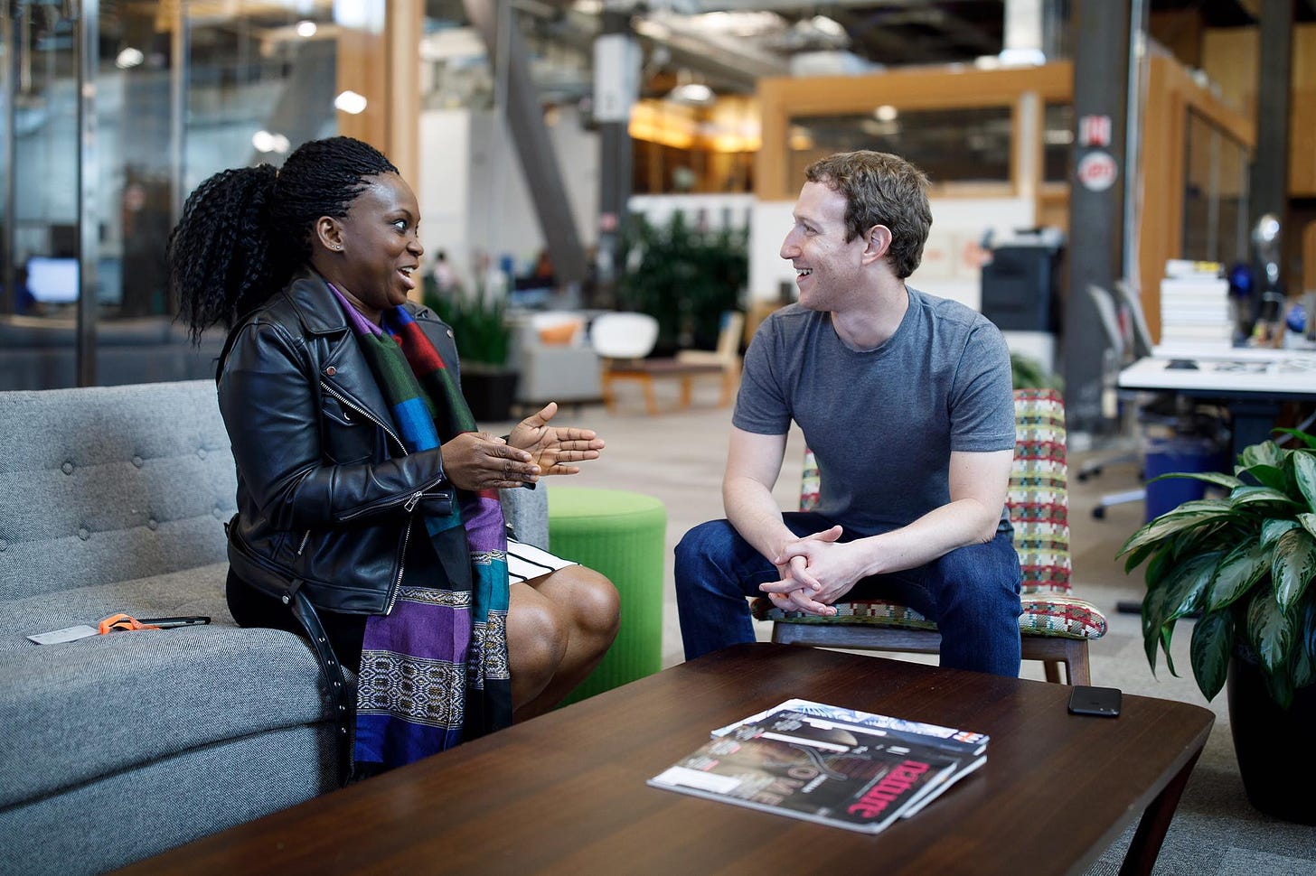 A photo of two people sitting and talking in a fancy office. At left, Lola is a Black woman with large braids wearing a leather jacket and long colorful scarf. At right is Mark Zuckerberg, a white man with jeans and a gray t-shirt.