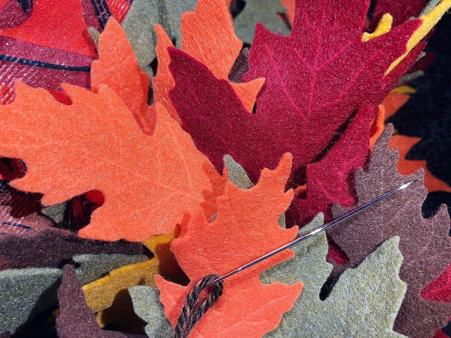 A photo of autumn colored felt leaves in orange, red, brown, gold, and green. On top of them is a large, shiny silver needle with brown yarn through it.