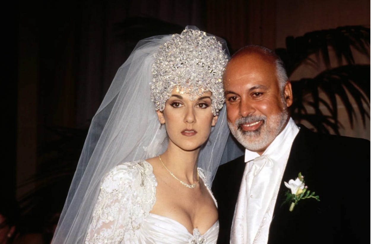 Celine Dion stands next to her husband, René Angélil, on her wedding day in 1994. She wears her white wedding dress and an elaborate silver headpiece with a veil and her spouse wears a black suit with a white flower pinned to his jacket.