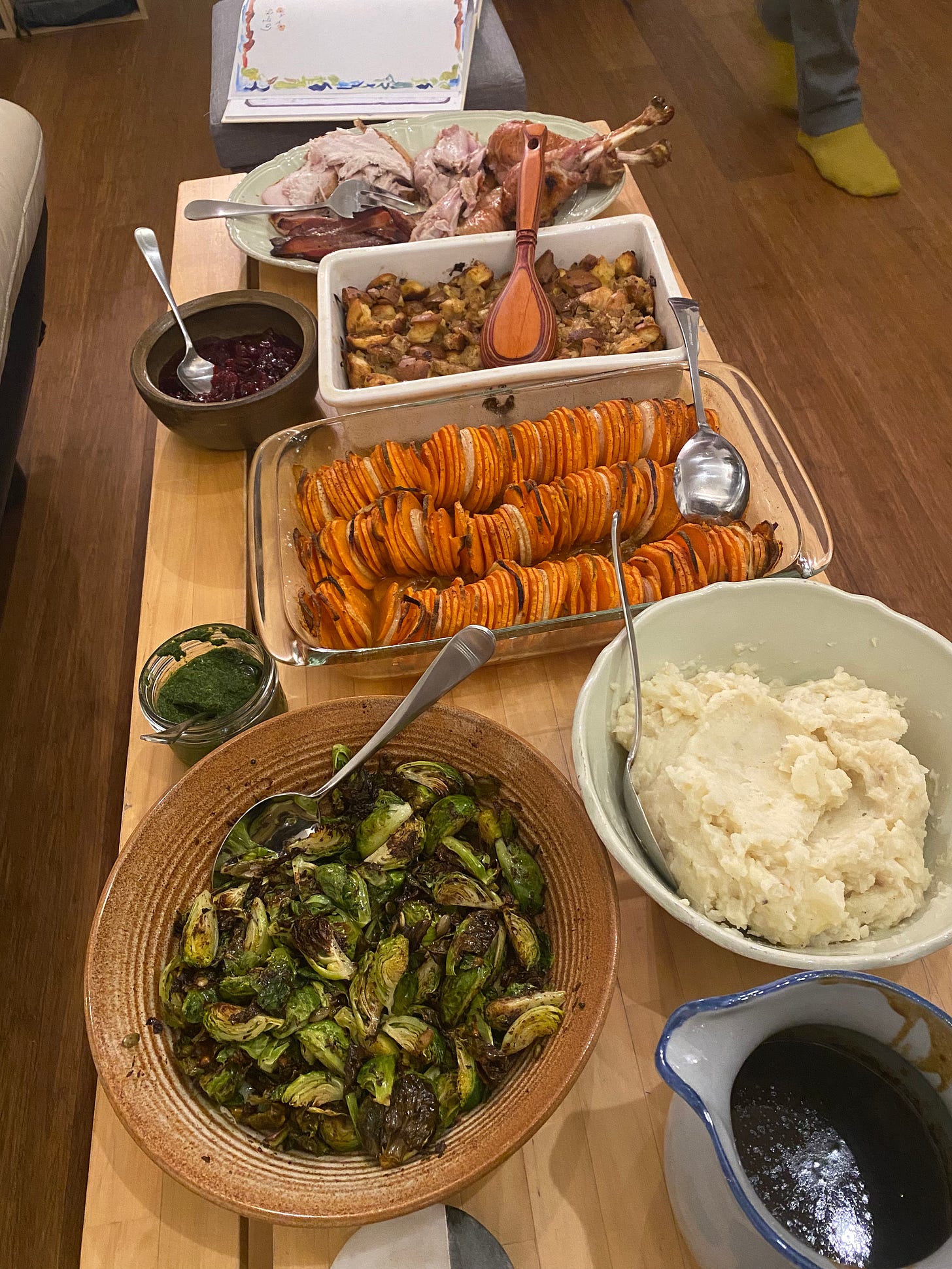 The coffee table, spread with thanksgiving dishes. From back to front: turkey, carved and on a green platter with a large serving fork. Stuffing in a white baking dish. Cranberry sauce in a small brown ceramic. A glass roasting dish with thinly sliced roasted yams, a small dish of chermoula next to it. A green bowl (matches the platter) of mashed potatoes & celery root. An orange-brown, wide bowl of roasted brussels sprouts. Gravy in a blue and grey pitcher.