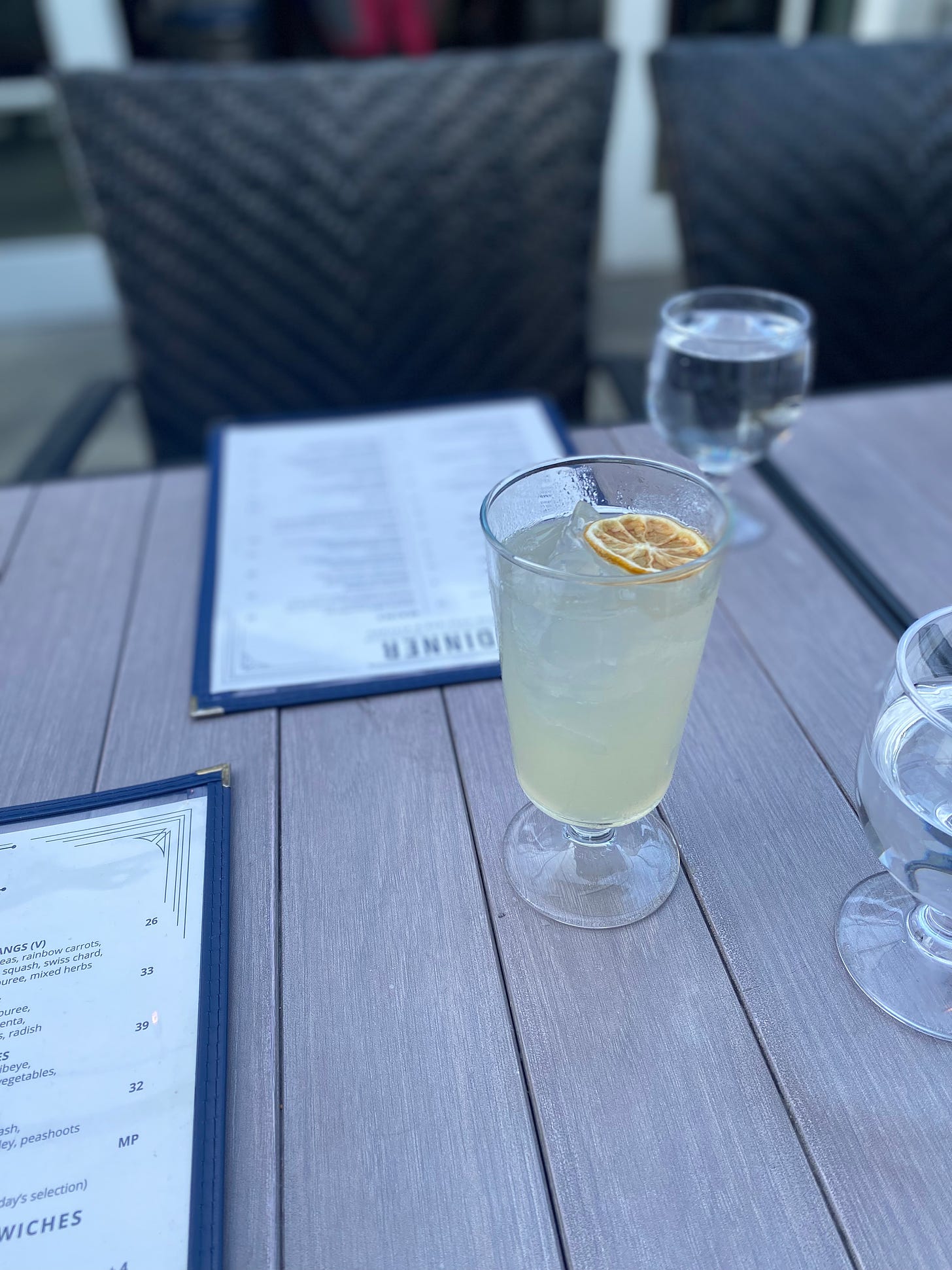 A patio table with two blue and white menus in front of each seat. Near the centre of the frame is a yellowish cocktail, the Gull's St George Soda, with a dried lemon floating on top of the ice.