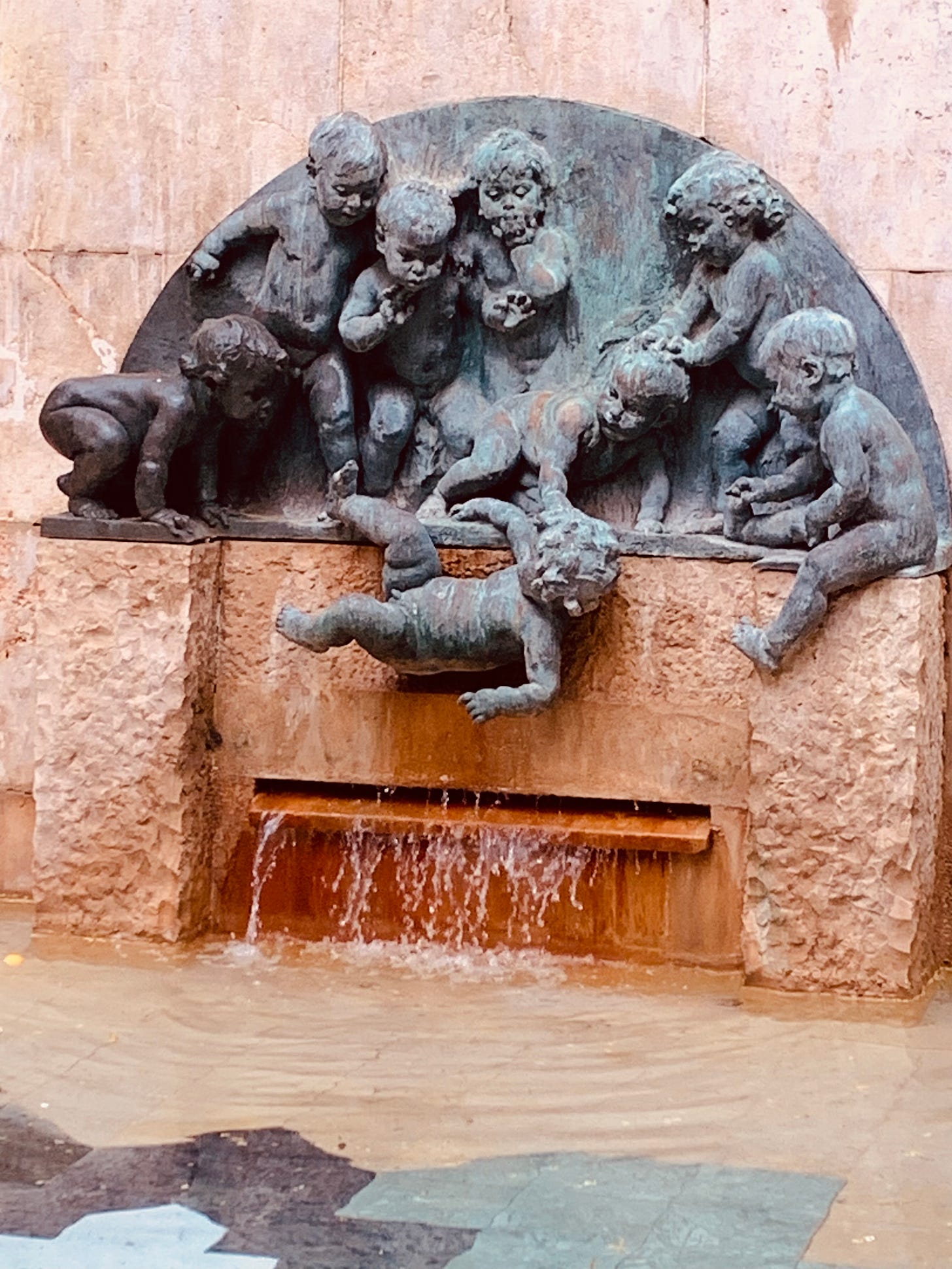 Above a fountain, a bas relief of a group of toddlers watches as one of their group falls off the ledge to the pool below.