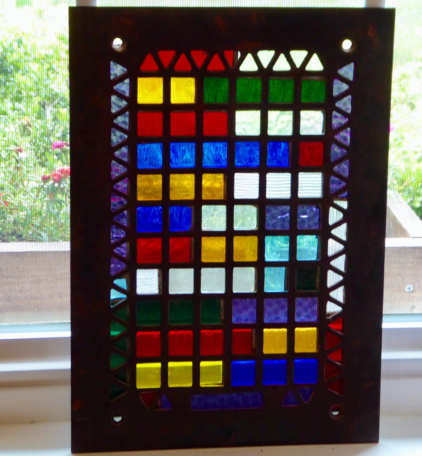 An old rectangular decorative heating grate with brightly colored squares of stained glass behind it.