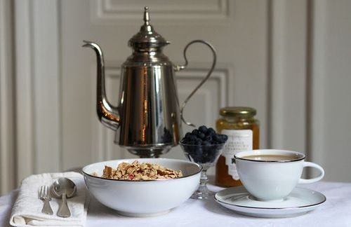 This may contain: a bowl of granola next to a cup of coffee on a saucer and spoon