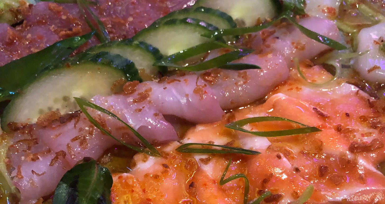 Detail from a plate of sushi featuring rows of slices of raw fish and sliced cucumber with a red and green garnish.