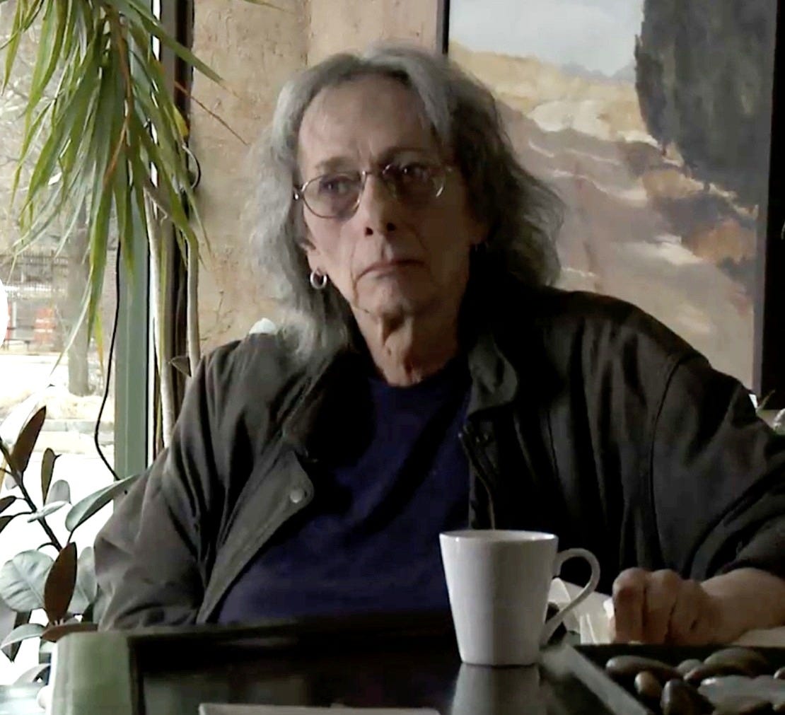 Color still of Jeffrey Catherine Jones late in life, seated at a café table with a coffee cup and stones on a tray. She wears a dark jacket and glasses, with long gray hair, gazing solemnly past the camera. A painting and houseplants frame the background. Color still of Jeffrey Catherine Jones late in life, seated at a café table with a coffee cup and stones on a tray. She wears a dark jacket and glasses, with long gray hair, gazing solemnly past the camera. A painting and houseplants frame the background.