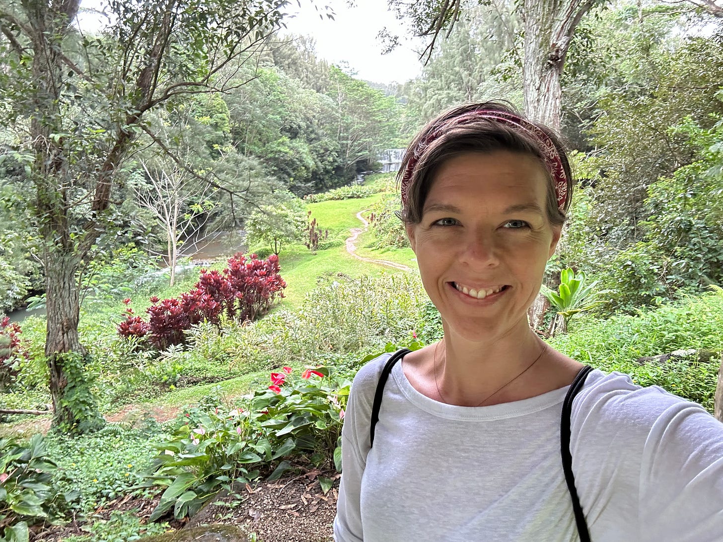 Woman with short brown hair and a white shirt takes a selfie in front of a waterfall and ravine full of tropical plants in Hawaii.