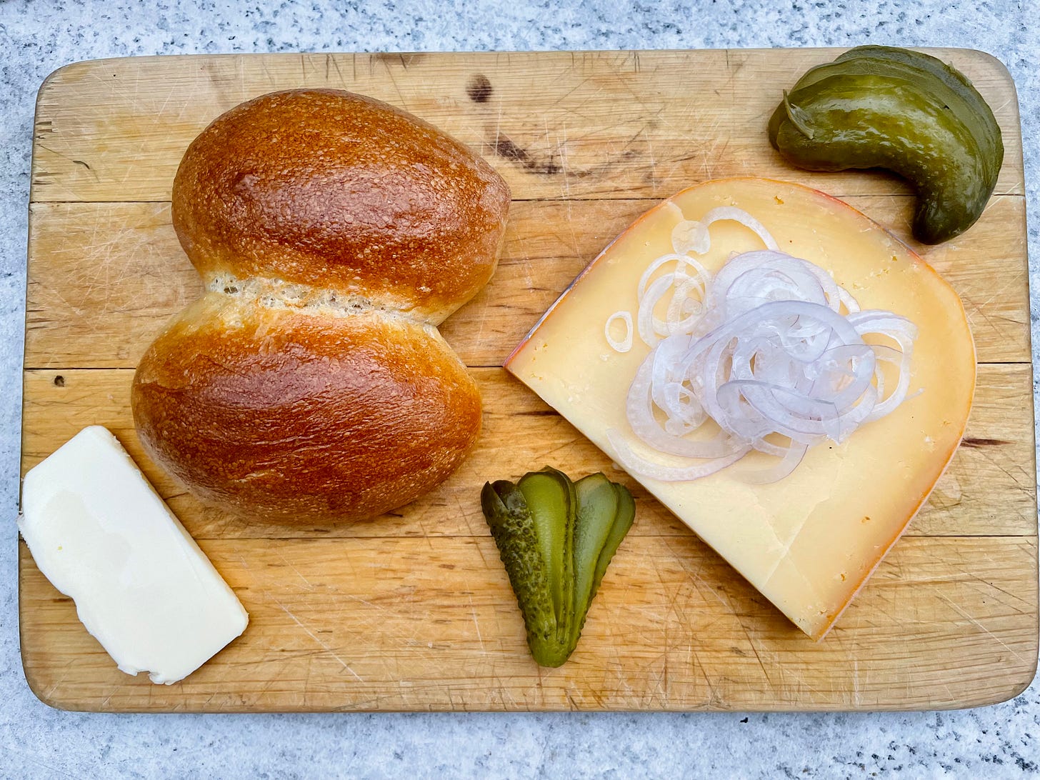 "A wooden board with the components of a Halve Hahn: a Röggelchen bun, a thick slice of aged Gouda cheese topped with thinly sliced onions, two types of pickles, and a piece of butter. A savory snack that can be enjoyed without beer." "A wooden board with the components of a Halve Hahn: a Röggelchen bun, a thick slice of aged Gouda cheese topped with thinly sliced onions, two types of pickles, and a piece of butter. A savory snack that can be enjoyed without beer."