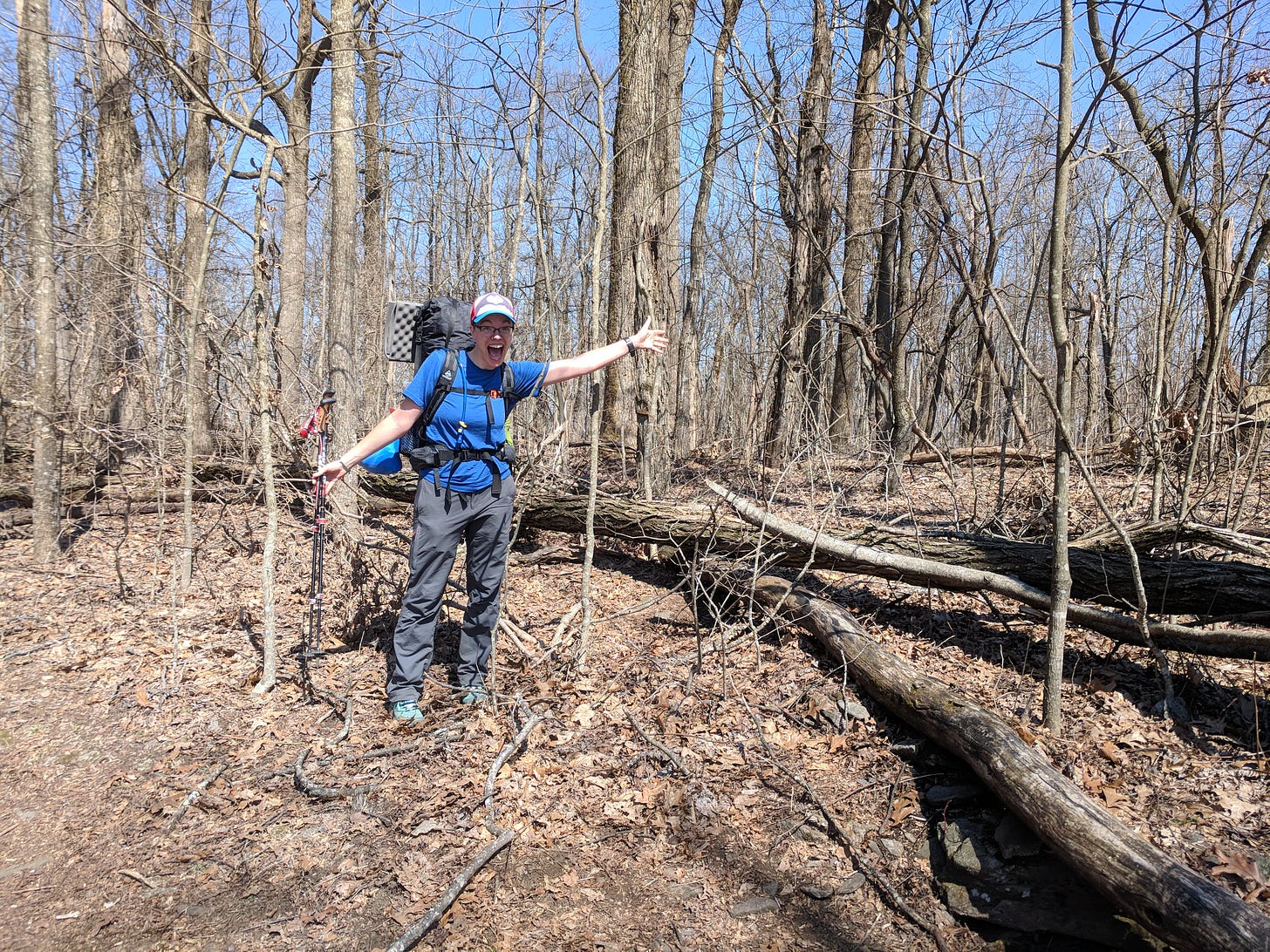 A photo of Kate on the Appalachian Trail. She wears a black backpack with a grey sleeping pad strapped to the top, carries two hiking poles, and has her arms spread wide and a big smile on her face. The scenery is boring: dead trees and trees devoid of leaves, but this is the summit of Sassafras Mountain in Georgia.