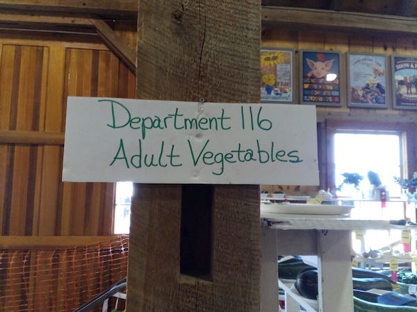 Crudely hand-lettered sign affixed to a rough post inside a large barn: "Department 116 Adult Vegetables"