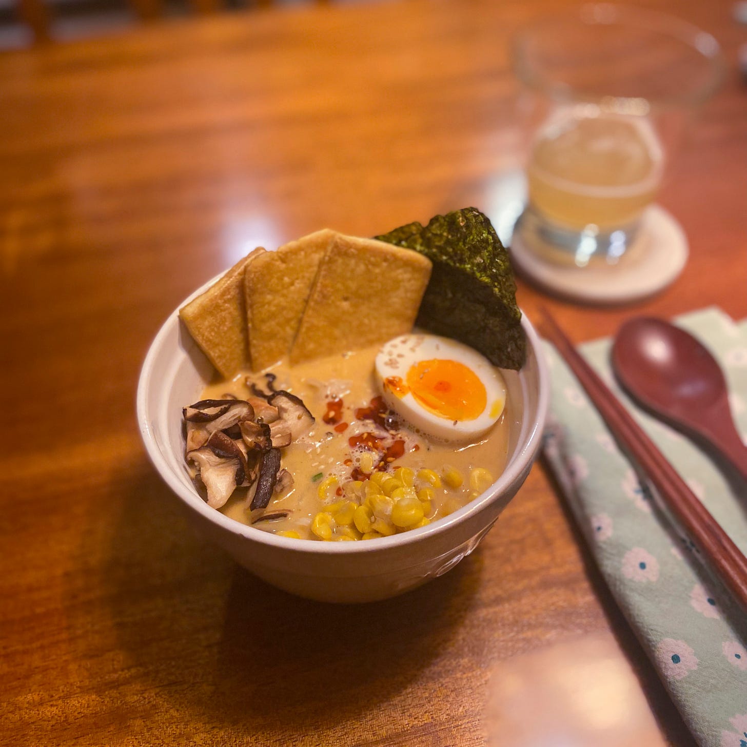 A white bowl of ramen with slices of fried tofu, shiitake mushrooms, corn, half a boiled egg, and a sheet of seaweed. Sesame seeds and chili oil are sprinkled on top. Next to it on a green flowered napkin are a set of wooden chopsticks and a matching spoon.