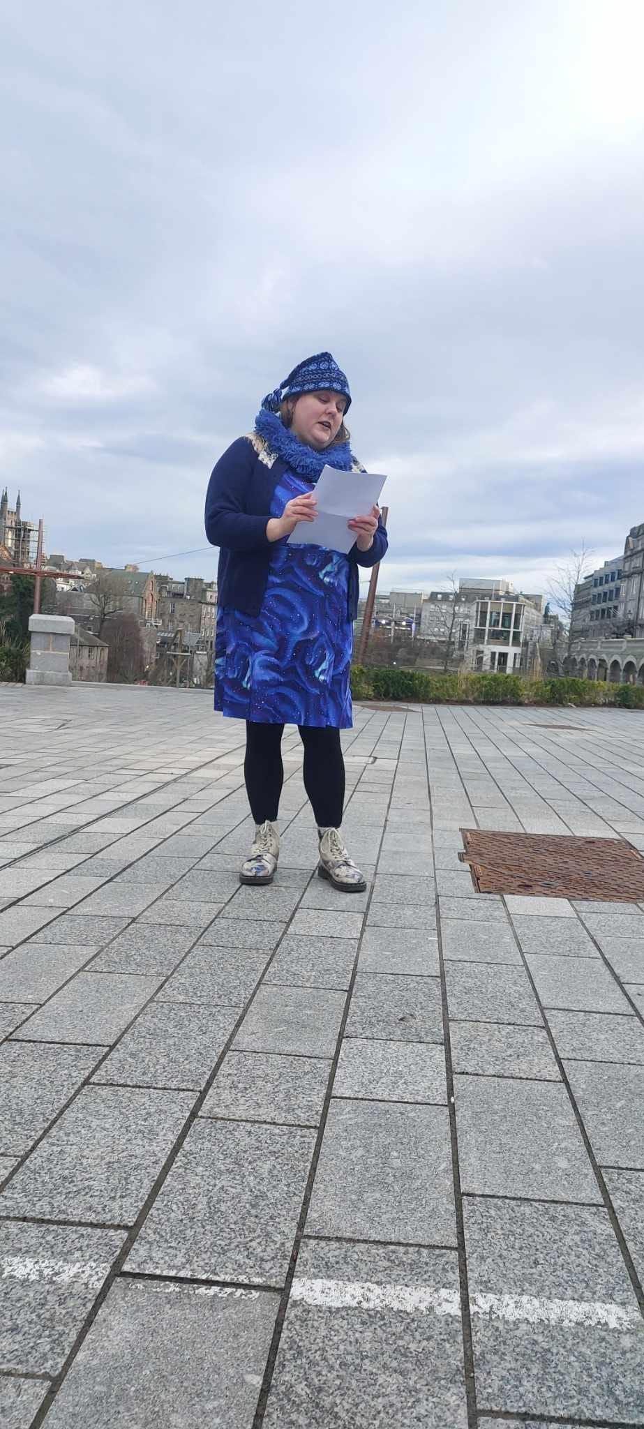 Hannah, a 35-year-old white female with blonde hair and blue eyes, is wearing a blue Fair Isle toorie, a navy Fair Isle yoke cardigan, a blue Popsy dress with a northern lights pattern, black Merino wool tights, and a pair of Dr Martens boots with a Great Wave of Kanagawa design. She is standing in Union Terrace Gardens in Aberdeen reading poetry from sheets of paper. 