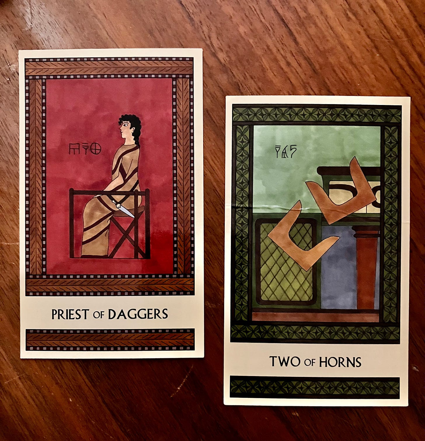 Two Minoan Tarot cards side by side on a brown wood surface. The Priest of Daggers is in shades of dark red and tan. It shows a Minoan man in a long robe, seated, facing left, with a dagger on his belt. The Two of Horns is in shades of green. It shows two pairs of sacred horns tumbling off a shrine.