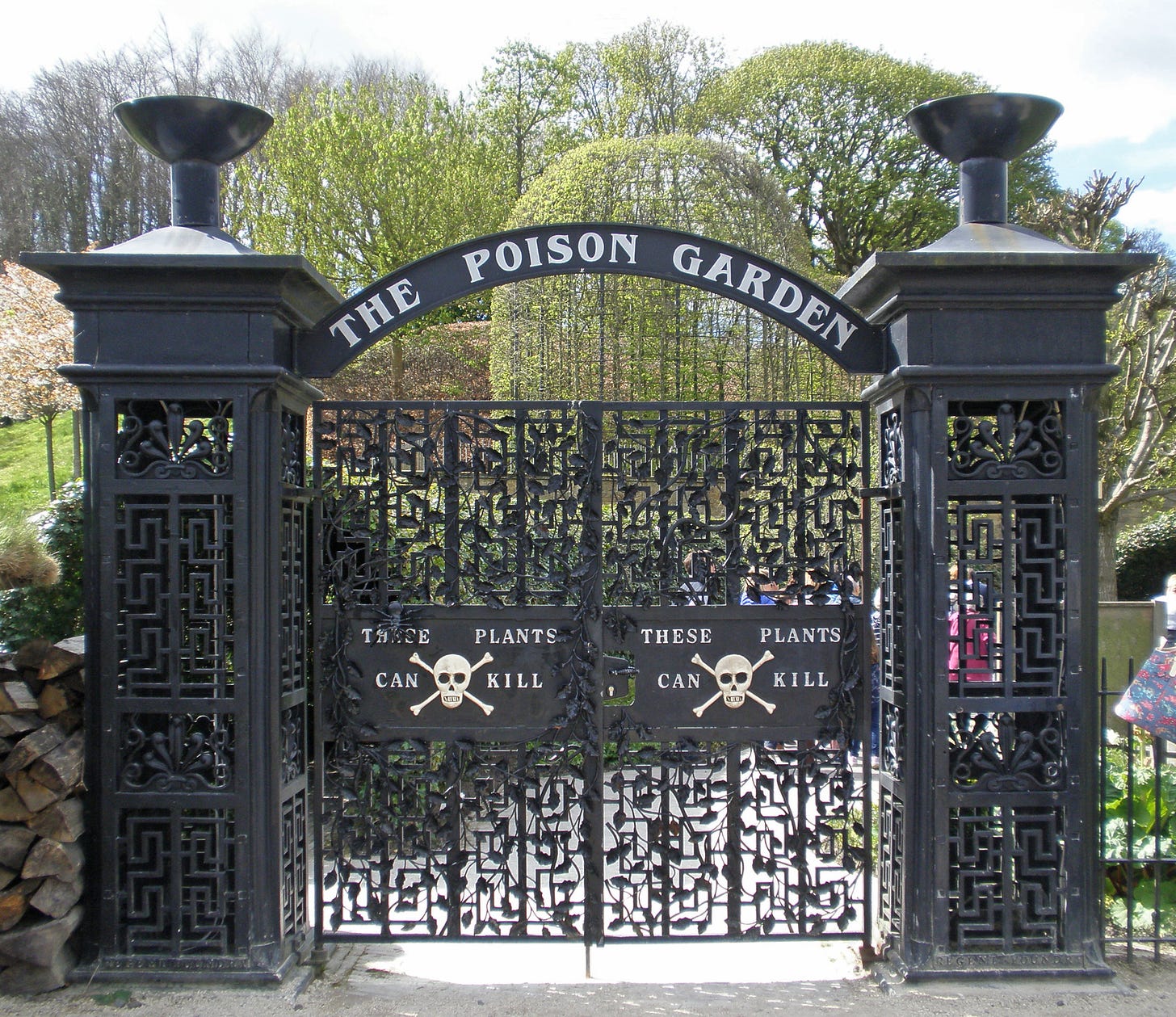Black geometric gates, with signs that have a skull and crossbones on them stating these plants can kill. Behind the gates, a very green garden can be glimpsed
