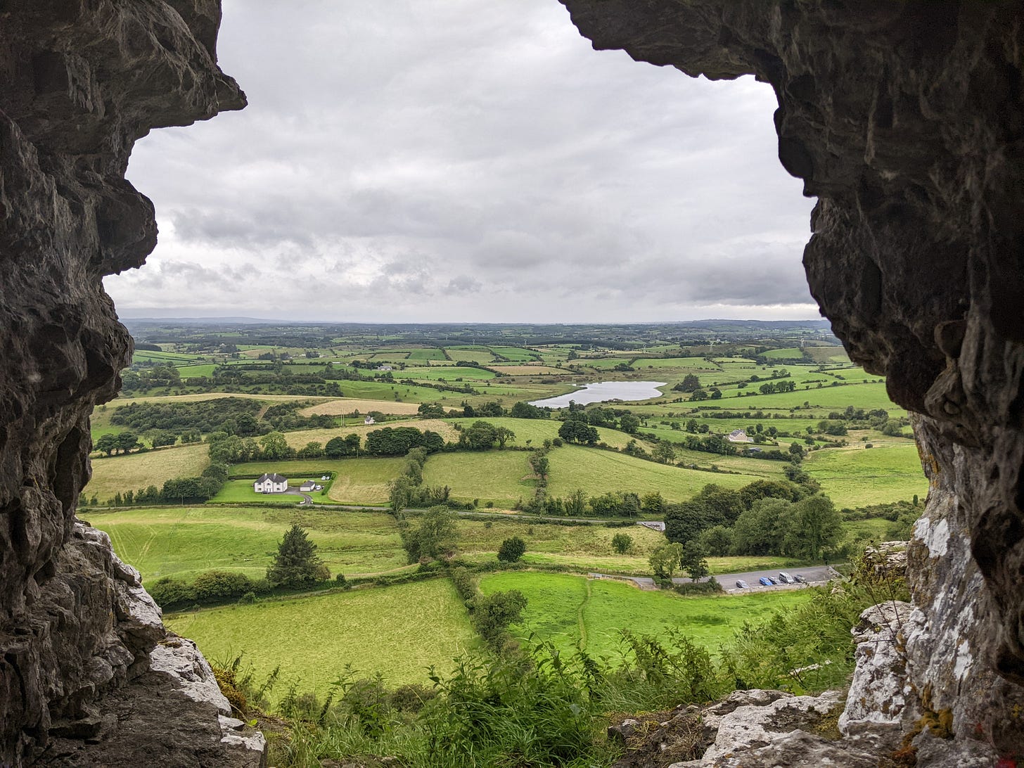 Framed by cave walls, a view out into the rolling landscape with hedges and trees and a slate-grey sky