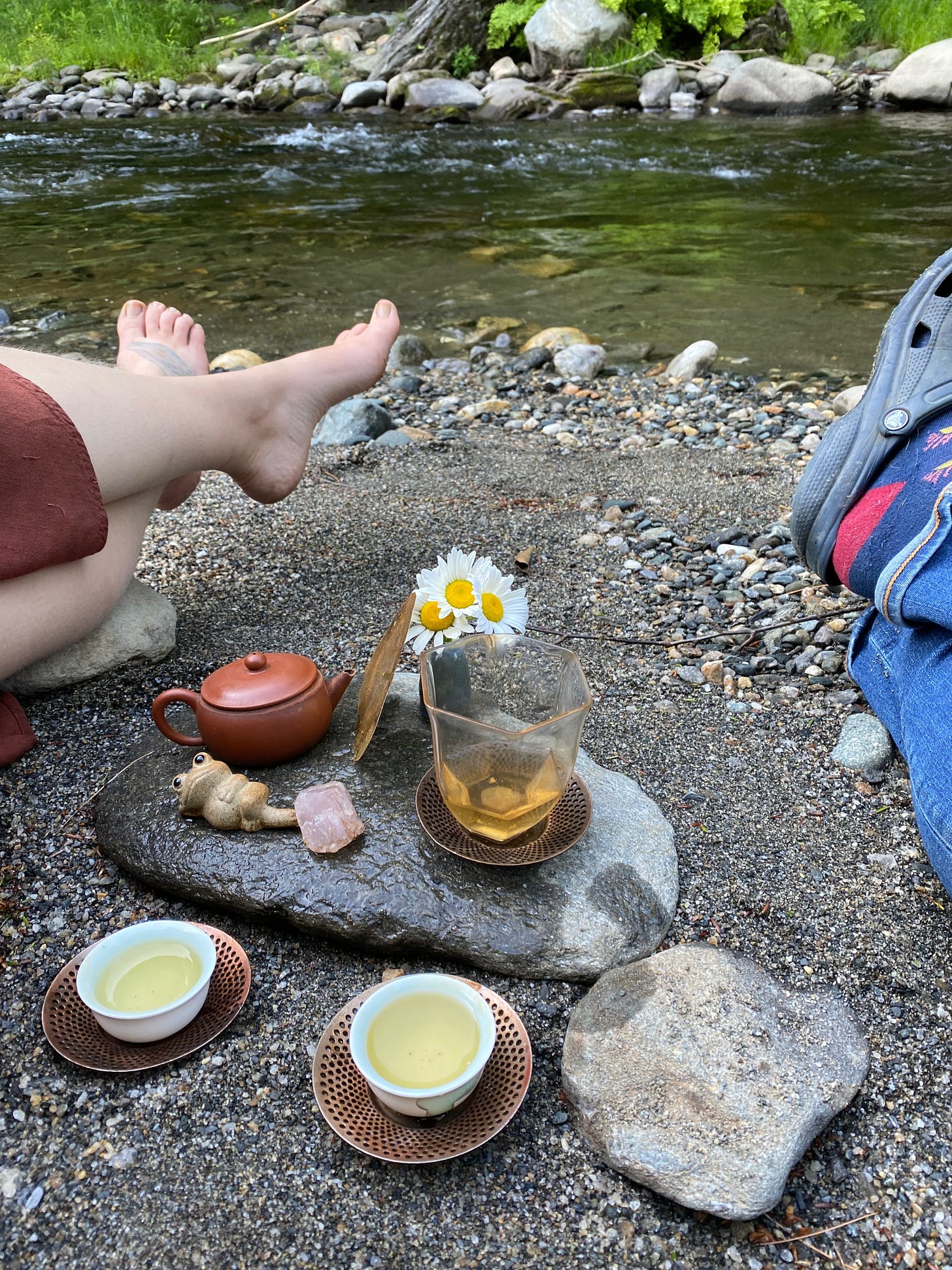 A tea setup on a flat rock by a river: two small cups on bronze saucers, a small teapot, a frog tea animal, a tiny vase of daises. The crossed, outstretched legs of two people are visible on either side of the tea setup.