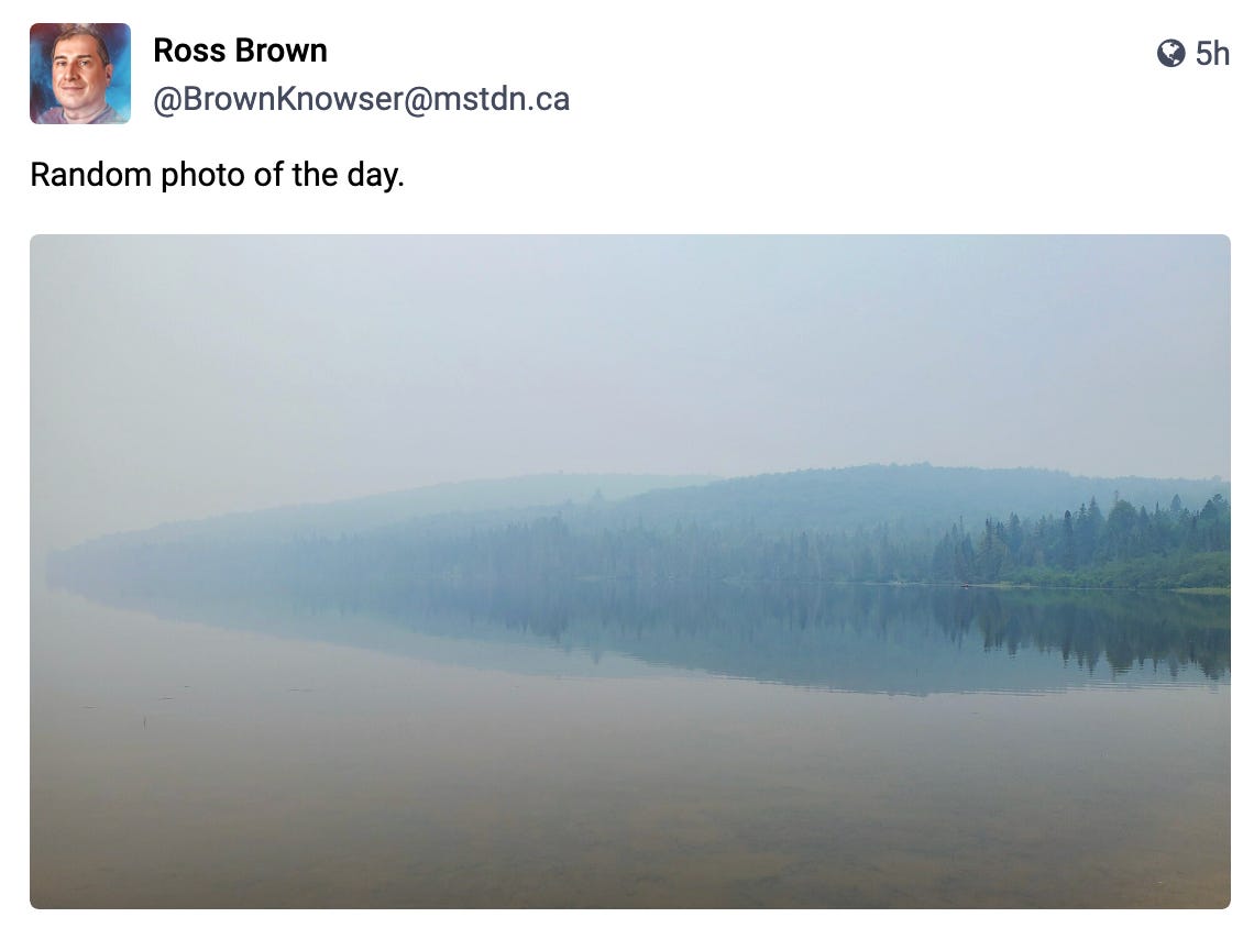The reflective water of Rock Lake in Algonquin Provincial Park is obscured by heavy smoke from distant wildfires. Low, rolling forested hills are captured in a haze on the far side of the lake.