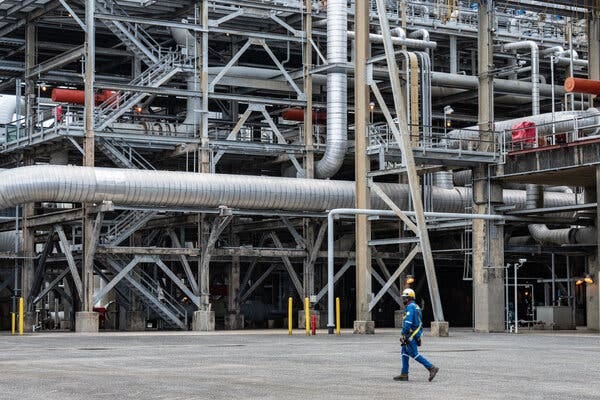 A worker in a blue uniform and a hard hat walks in front of a liquefied natural gas export terminal.