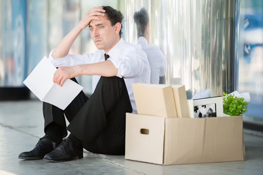 A man sits on the ground with his belongings in a box after losing his job, looking distressed and uncertain about the future.