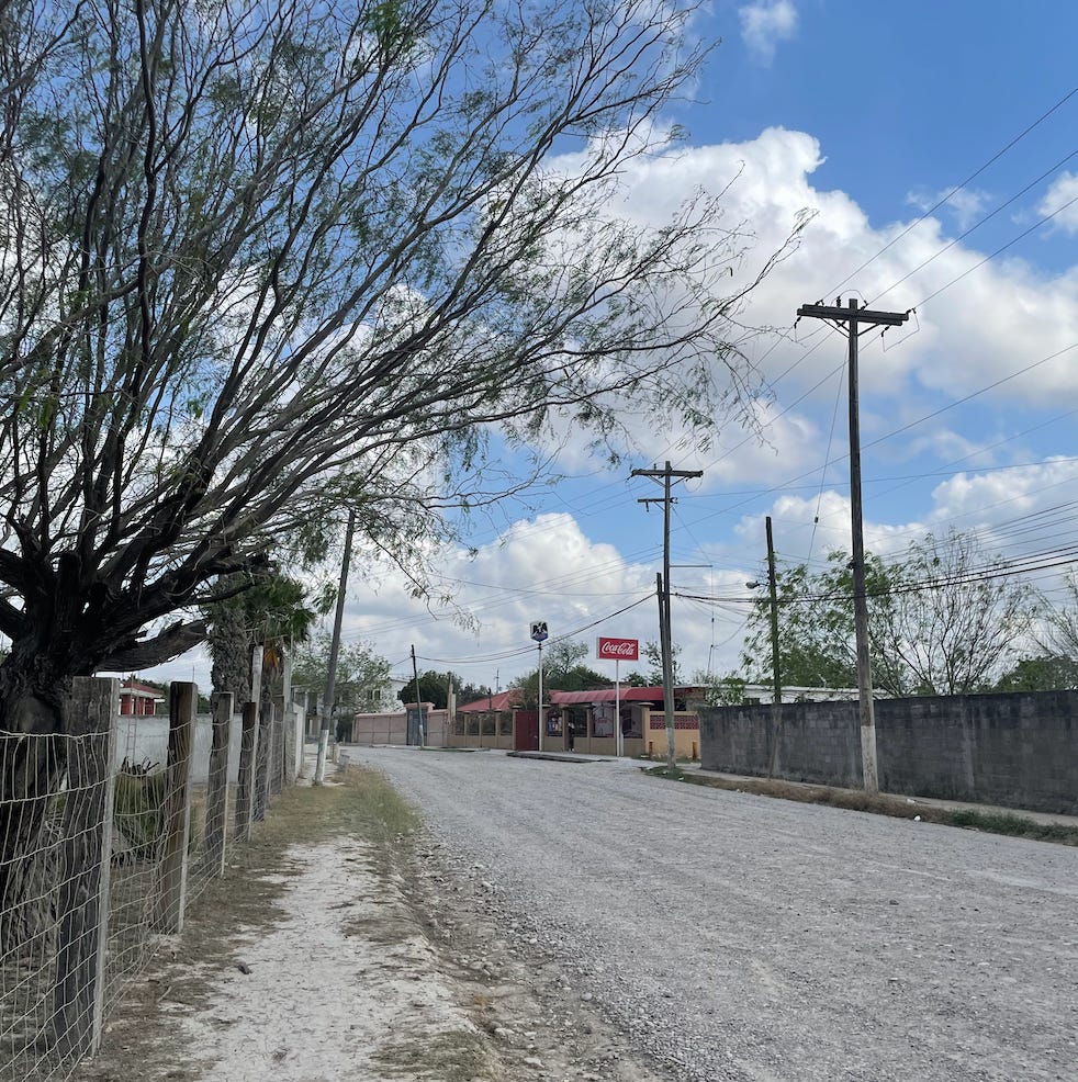A photograph of a rocky dirt road in a rural Mexican ejido. Mesquite trees line the road. Across the road, you can see a Coca-Cola sign and a Tecate sign. The right side of the road is lined with electrical poles. The sky is a light blue with many cumulus clouds.