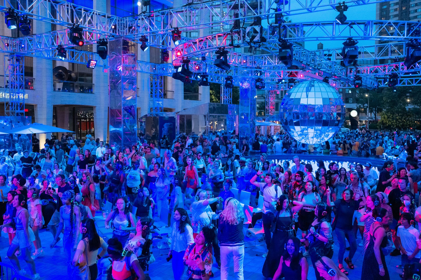 A photo of a huge outdoor dance floor with people wearing headsets and haptic suits, bathed in blue. A 10 foot tall disco ball spins over bubbling fountains.